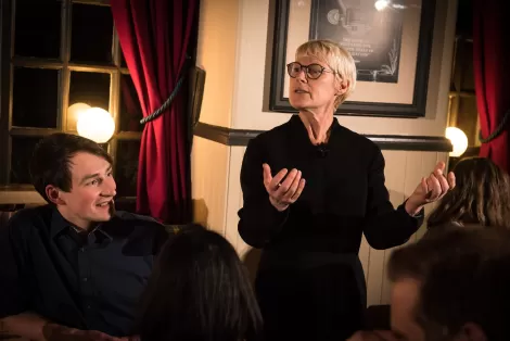 Woman standing and gesticulating whilst telling a story to a group of people in a pub at night