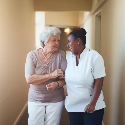 Female care worker assisting an elderly woman down a bright corridor