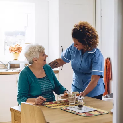 Female nurse stands above an elderly woman sat at the kitchen table with her hand on her shoulder, both smiling at each other